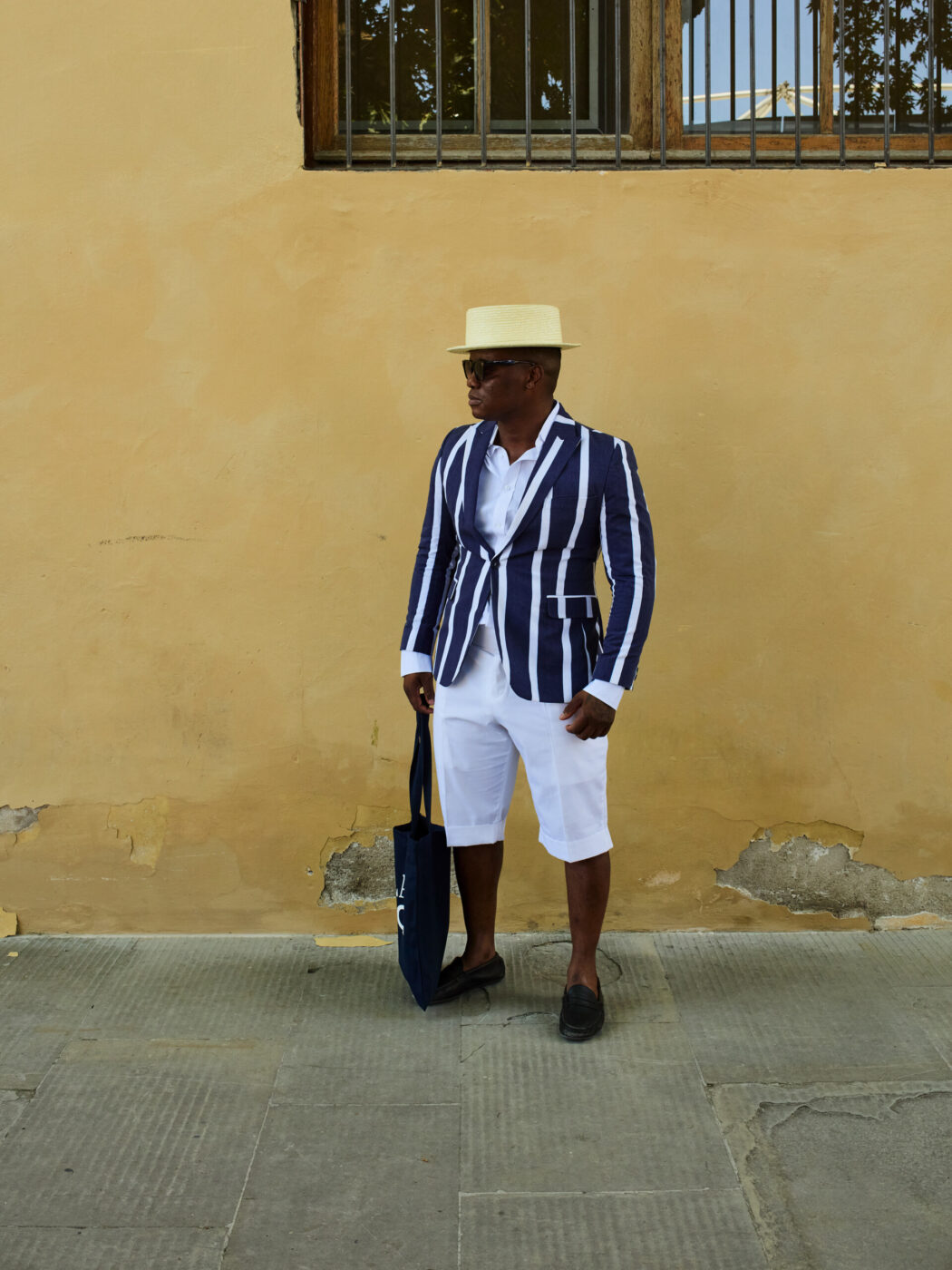 [Un uomo che indossa un cappello color crema, occhiali da sole, un blazer a righe bianche e blu, pantaloncini bianchi e mocassini scuri è in piedi contro un muro giallo e tiene al fianco una borsa blu.