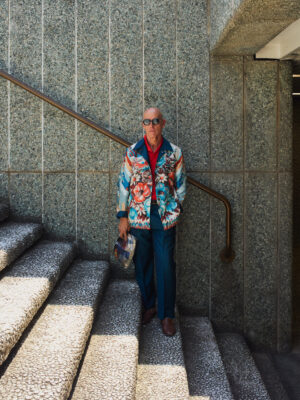 An older man in sunglasses stands on stone stairs against a textured wall, wearing a colorful floral jacket, blue pants, brown shoes, and holding a patterned bag. Sunlight highlights part of the stairs. If you add an image title (e.g., Fashion Statement), the formatted alt text would be: Fashion Statement - An older man in sunglasses stands on stone stairs against a textured wall, wearing a colorful floral jacket, blue pants, brown shoes, and holding a patterned bag. Sunlight highlights part of the stairs.