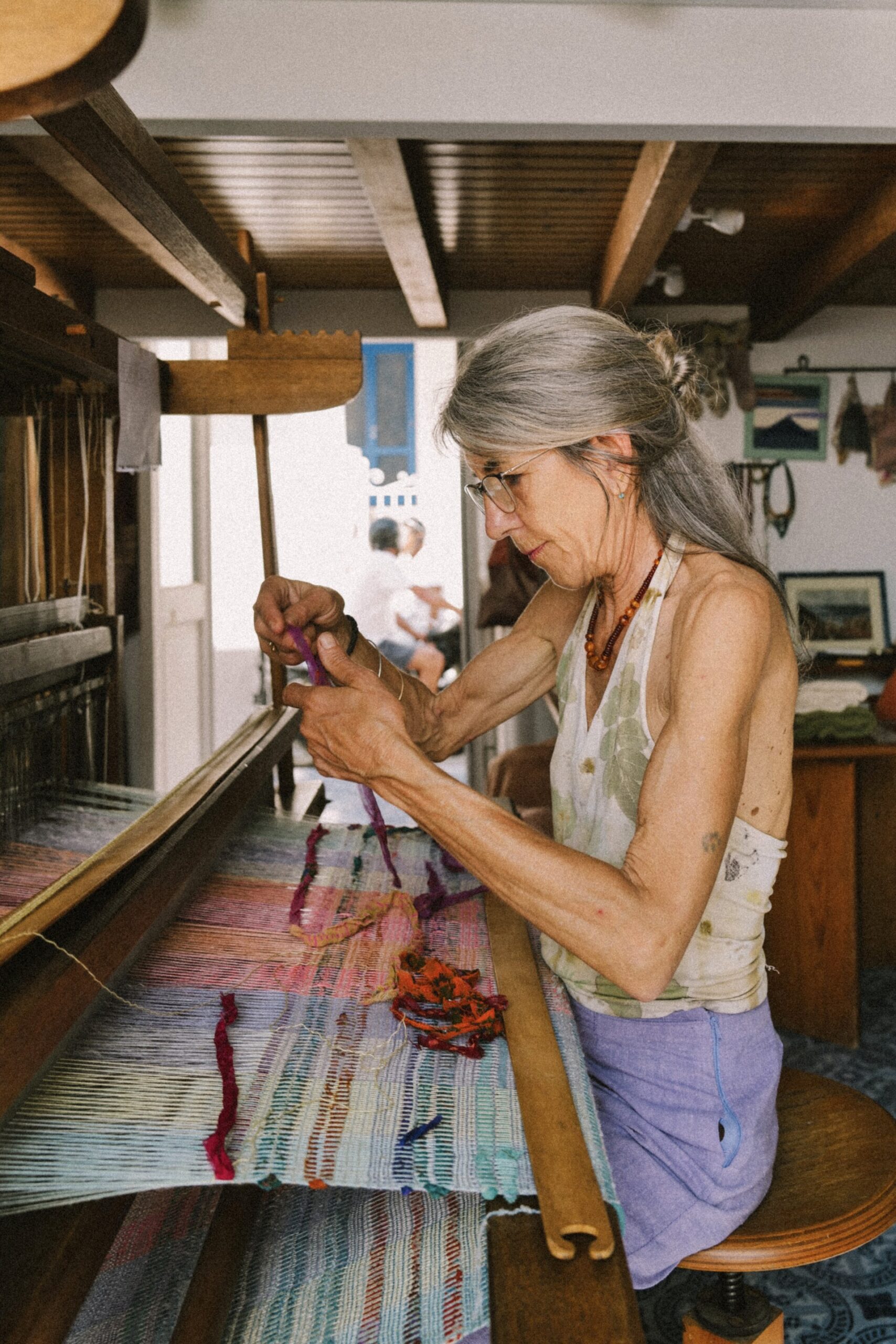 An older woman weaving on a loom - An older woman with long gray hair and glasses weaves colorful threads on a loom in a cozy, sunlit room filled with craft materials and decor.