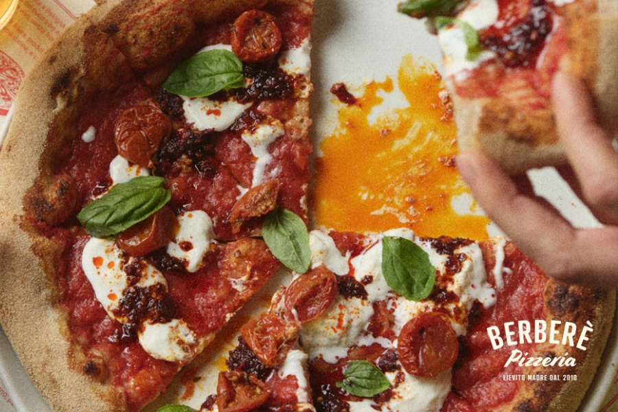 Berberè Pizzeria - A hand lifts a slice of pizza topped with tomato sauce, cherry tomatoes, mozzarella, basil leaves, and chili oil. The pizza is on a white plate with the text Berberè Pizzeria in the corner.