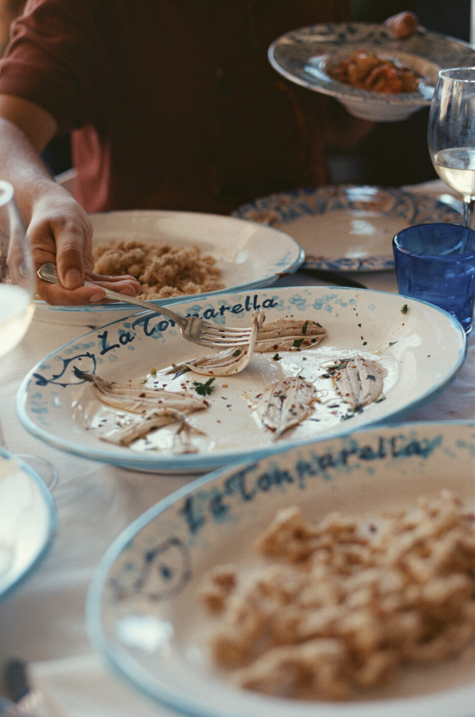 Una tavola apparecchiata con piatti di pesce parzialmente consumati - Una tavola apparecchiata con piatti di pesce parzialmente consumati su piatti bianchi e blu, con una persona che tiene un piatto e serve il cibo. Sono visibili anche bicchieri da vino e un bicchiere d'acqua blu.