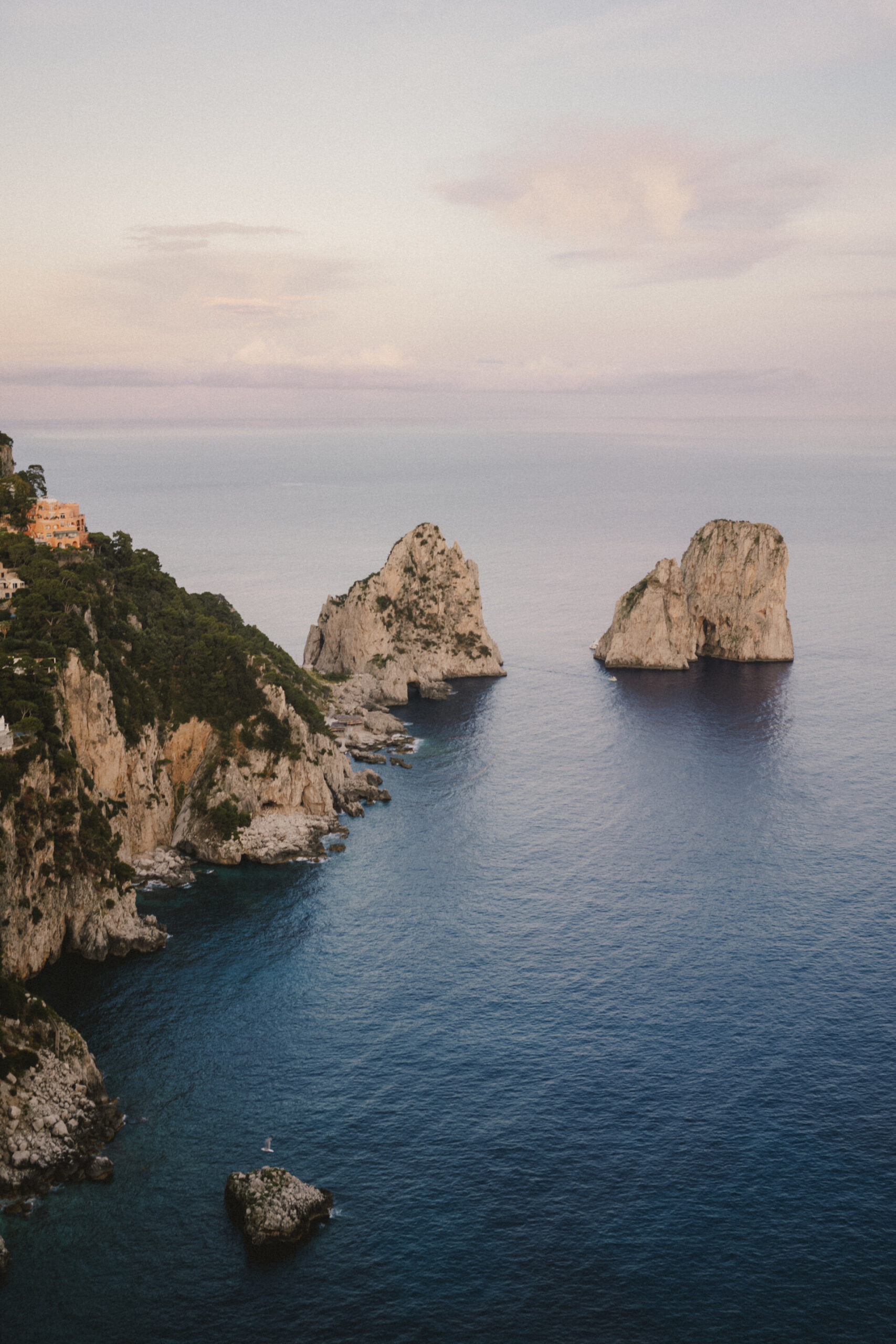 Formazioni rocciose al tramonto - Formazioni rocciose si ergono dal calmo mare blu vicino a una costa frastagliata con vegetazione ed edifici, sotto un cielo pastello al tramonto.