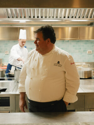 Uno chef in una cucina moderna - Uno chef in uniforme bianca sorride in una cucina moderna, con altri due chef che lavorano sullo sfondo vicino ai fornelli e alle attrezzature in acciaio inossidabile.
