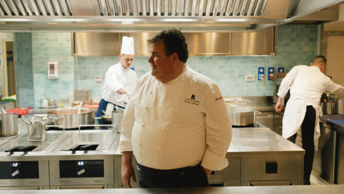 Uno chef in una cucina moderna - Uno chef in uniforme bianca sorride in una cucina moderna, con altri due chef che lavorano sullo sfondo vicino ai fornelli e alle attrezzature in acciaio inossidabile.