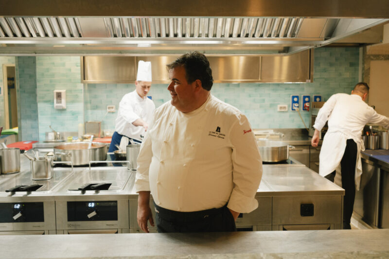 Uno chef in una cucina moderna - Uno chef in uniforme bianca sorride in una cucina moderna, con altri due chef che lavorano sullo sfondo vicino ai fornelli e alle attrezzature in acciaio inossidabile.