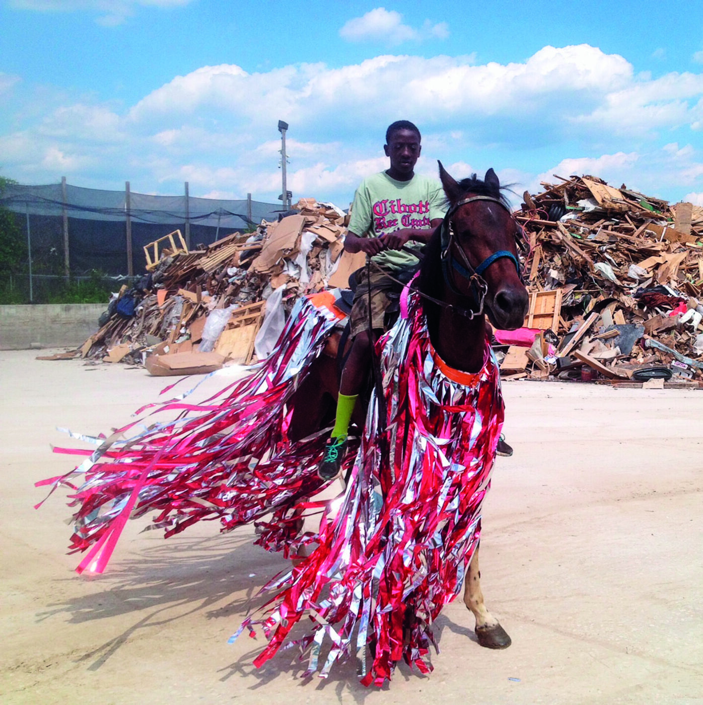 A person rides a horse decorated with long, colorful red, white, and silver streamers. Behind them is a large pile of debris and scrap materials under a blue sky with scattered clouds.