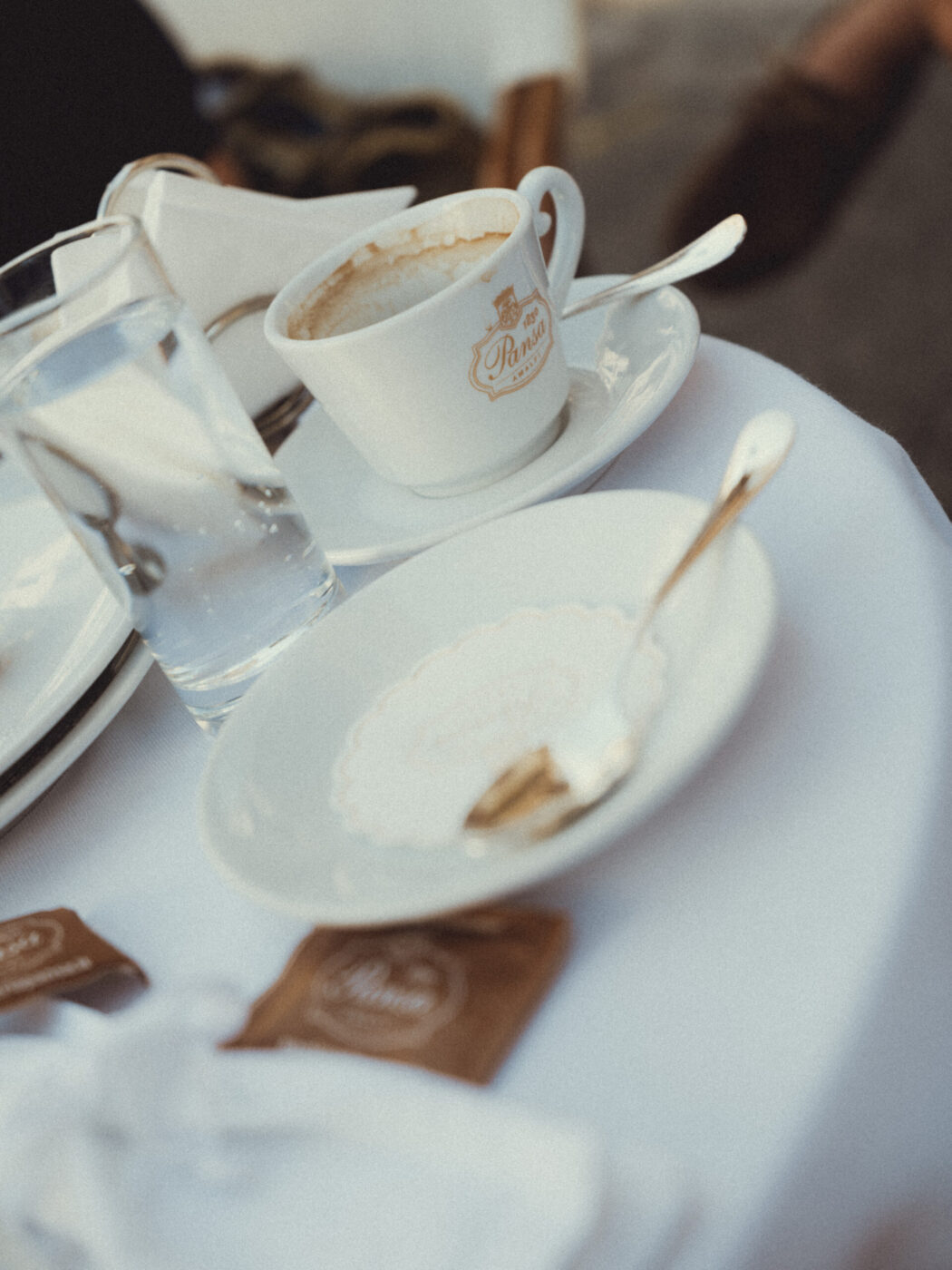Non è stato fornito alcun titolo per l'immagine. Ecco il testo alternativo formattato: Un tavolo bianco contiene una tazza di caffè, un bicchiere d'acqua, un piatto vuoto con un cucchiaio e due bustine di zucchero di canna. La scena sembra provenire da un bar all'aperto.