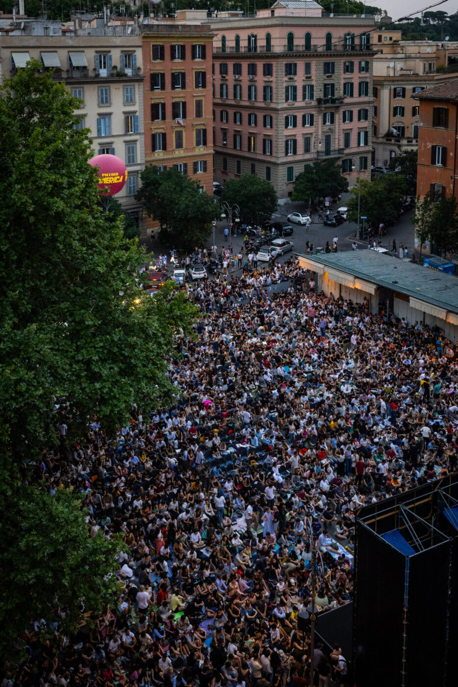 A large crowd fills an outdoor urban square - A large crowd fills an outdoor urban square, surrounded by tall buildings and trees, gathered for an event in the evening. The atmosphere appears lively and energetic.