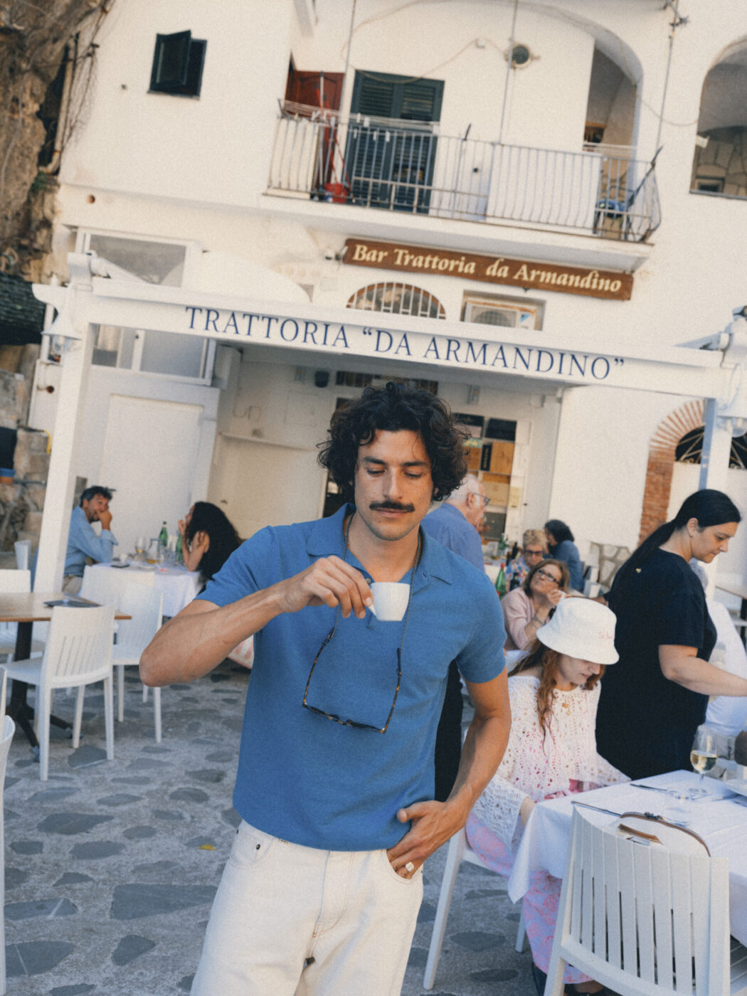 Trattoria da Armandino - Un uomo in camicia blu si trova all'esterno della Trattoria da Armandino, sorseggiando un caffè e tenendo in mano degli occhiali da sole. Intorno a lui le persone cenano ai tavoli bianchi, con un'atmosfera rilassata e soleggiata da caffè italiano sullo sfondo.