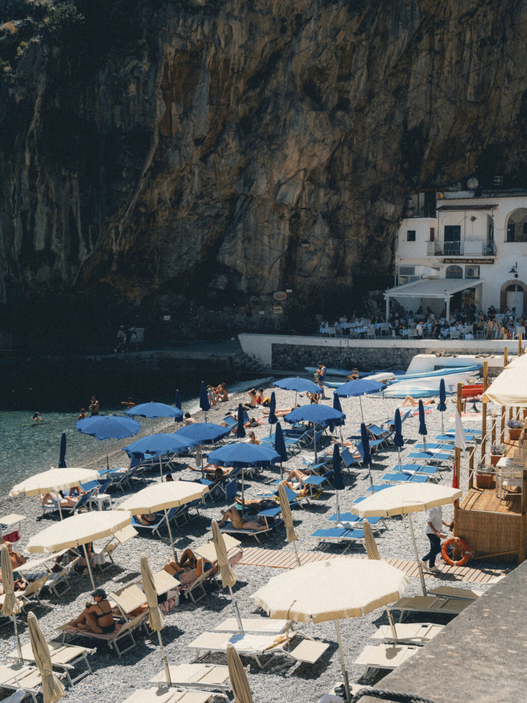 Scena di spiaggia affollata con file di ombrelloni e lettini blu e beige, persone che si rilassano e prendono il sole, accanto a calme acque verdi e a una grande scogliera rocciosa, con un edificio bianco sullo sfondo.