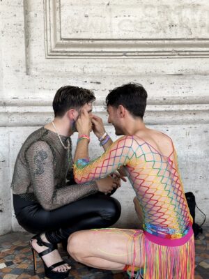 Due amiche - Due persone siedono l'una di fronte all'altra; una indossa un top nero a rete, pantaloni di pelle e tacchi, mentre l'altra indossa un abito a rete colorato. Quest'ultima trucca la prima, entrambe su un pavimento a mosaico contro un muro di pietra.