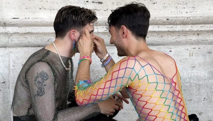 There is no image title provided. Alt text: Two men sit facing each other on a mosaic floor. One, wearing a rainbow fishnet dress, is applying makeup to the other, who is dressed in black mesh and high heels. They appear focused, with a stone wall behind them.