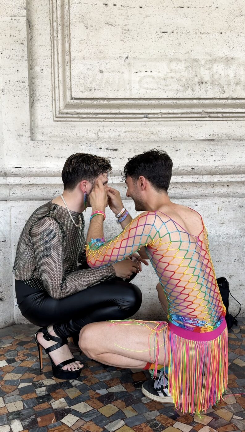 Due amiche - Due persone siedono l'una di fronte all'altra; una indossa un top nero a rete, pantaloni di pelle e tacchi, mentre l'altra indossa un abito a rete colorato. Quest'ultima trucca la prima, entrambe su un pavimento a mosaico contro un muro di pietra.