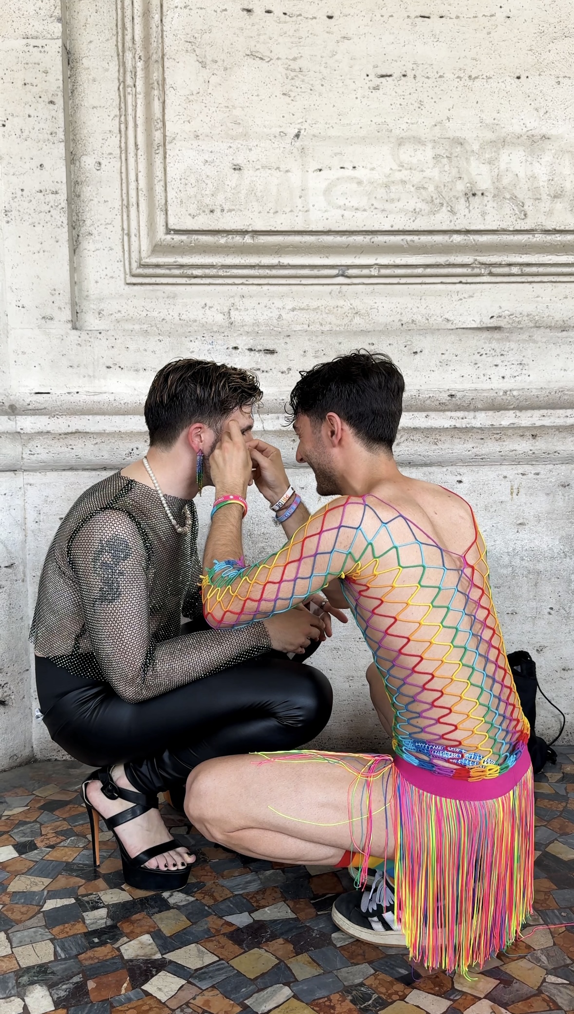 Due amiche - Due persone siedono l'una di fronte all'altra; una indossa un top nero a rete, pantaloni di pelle e tacchi, mentre l'altra indossa un abito a rete colorato. Quest'ultima trucca la prima, entrambe su un pavimento a mosaico contro un muro di pietra.