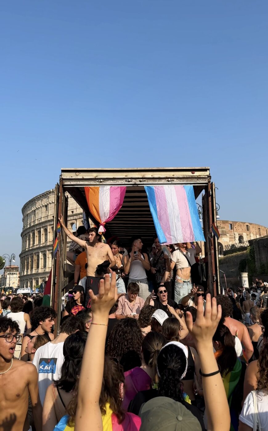 A lively crowd gathers near the Colosseum in Rome - A lively crowd gathers near the Colosseum in Rome. People dance atop a truck decorated with rainbow, lesbian, and transgender pride flags, celebrating under a clear blue sky. Raised hands show excitement and support.