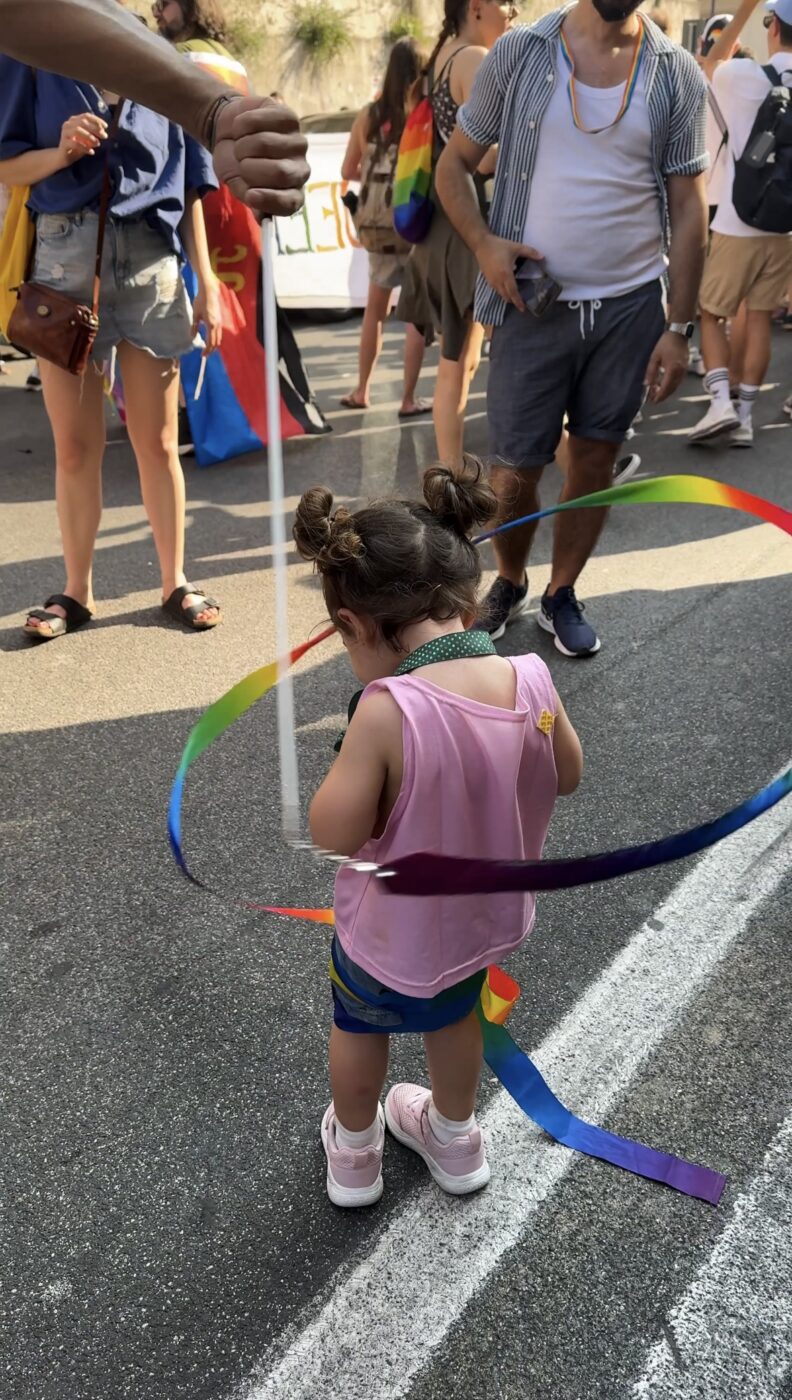 Un bambino con due chignon tra i capelli gioca con un nastro colorato durante un evento di strada. Adulti e altre persone sono in piedi, alcuni indossano o tengono in mano oggetti arcobaleno, suggerendo una celebrazione dell'orgoglio.