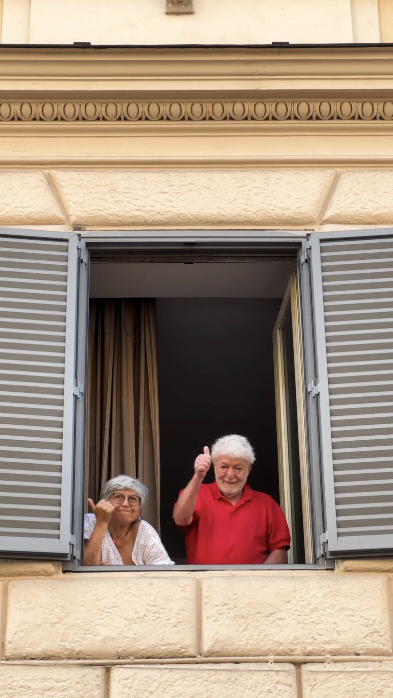 [Due adulti anziani, una donna e un uomo, sono in piedi davanti a una finestra aperta con le persiane grigie. L'uomo alza il pollice, mentre la donna saluta. Entrambi sorridono e l'edificio ha un esterno color crema con decorazioni in pietra.