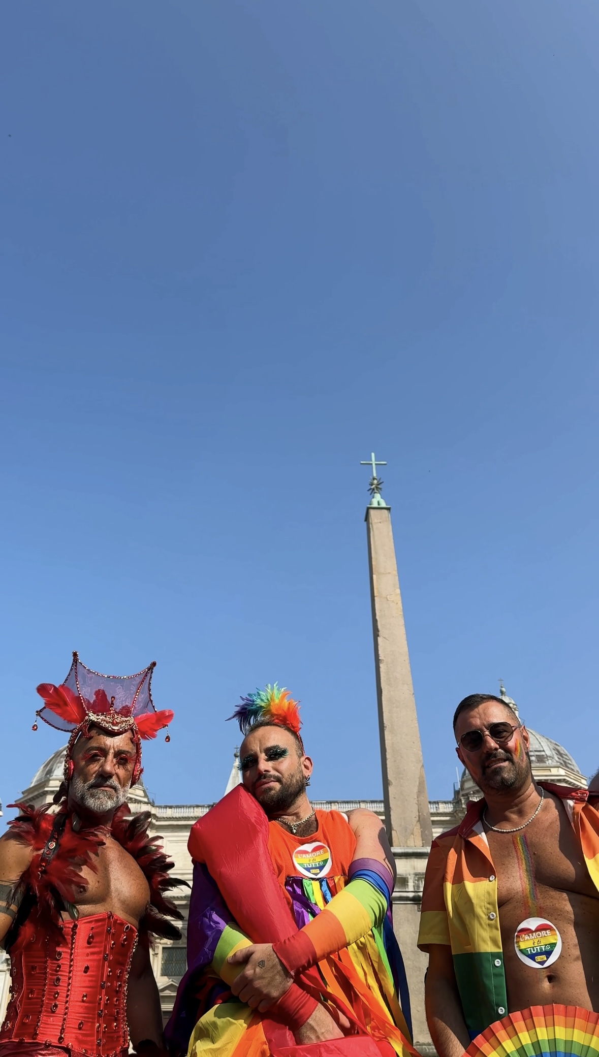 Tre uomini in abiti colorati e festosi, tra cui accessori arcobaleno e un copricapo di piume rosse, si trovano di fronte a un obelisco sotto un cielo azzurro e limpido, per festeggiare un evento pride.