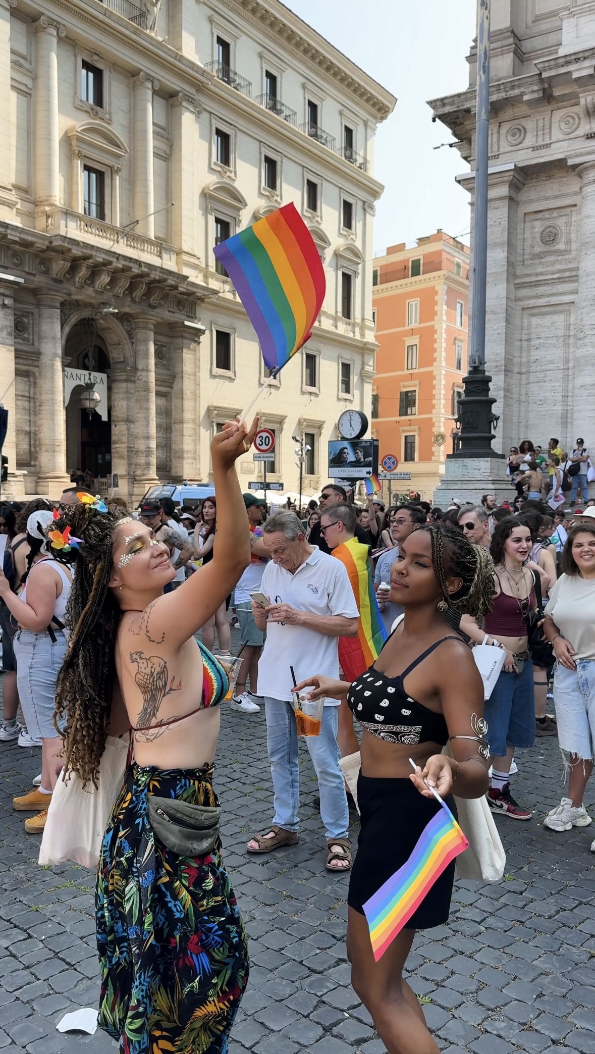 Una folla si raduna all'aperto per un evento Pride - Due donne in primo piano, una delle quali sventola una bandiera arcobaleno e sorride. Le persone intorno a loro appaiono gioiose, con colori arcobaleno e abiti festosi. Sullo sfondo, edifici storici.