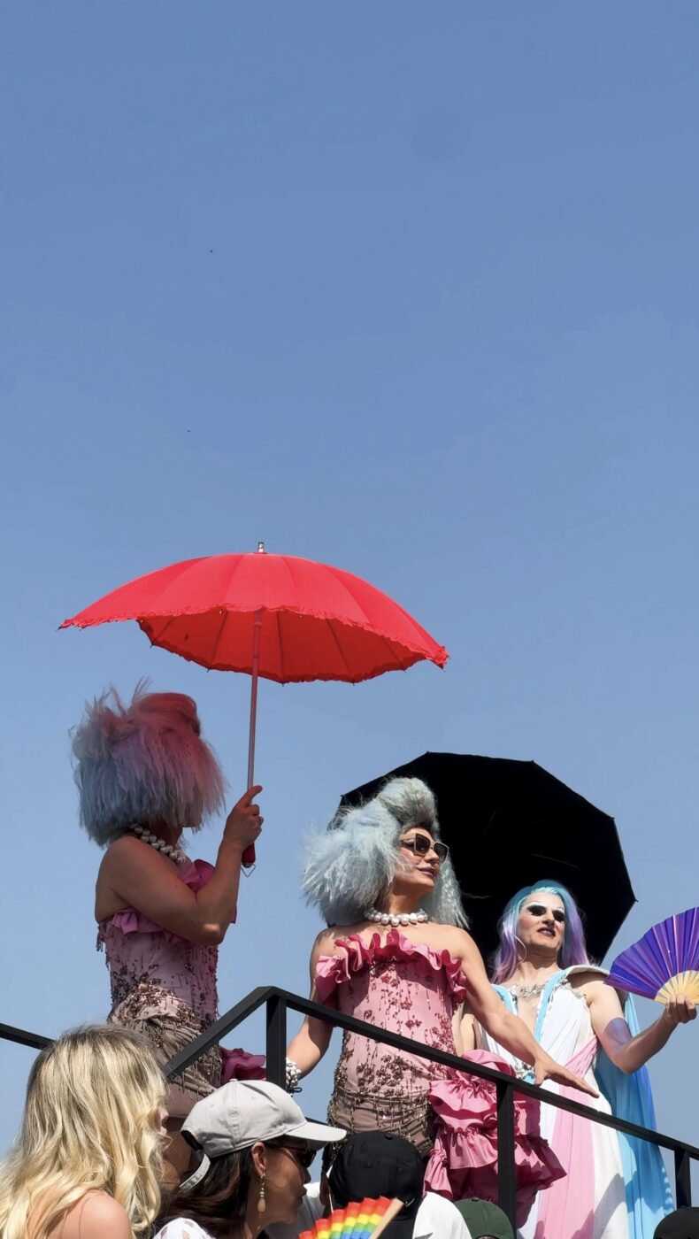 Tre persone con parrucche e occhiali da sole color pastello - Tre persone con parrucche e occhiali da sole color pastello si trovano su una piattaforma sotto un cielo azzurro, con ombrelli rossi, neri e viola in mano. Sono vestiti con abiti vivaci, probabilmente partecipano a un evento festivo all'aperto.