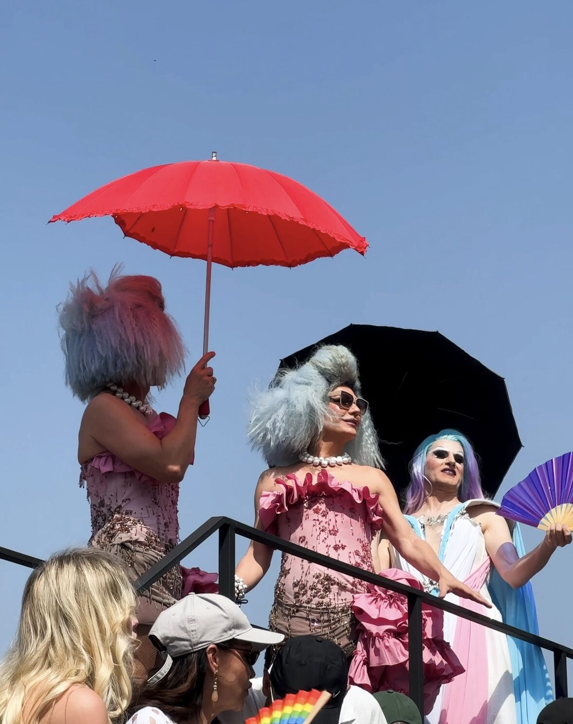 Tre persone con parrucche e occhiali da sole color pastello - Tre persone con parrucche e occhiali da sole color pastello si trovano su una piattaforma sotto un cielo azzurro, con ombrelli rossi, neri e viola in mano. Sono vestiti con abiti vivaci, probabilmente partecipano a un evento festivo all'aperto.