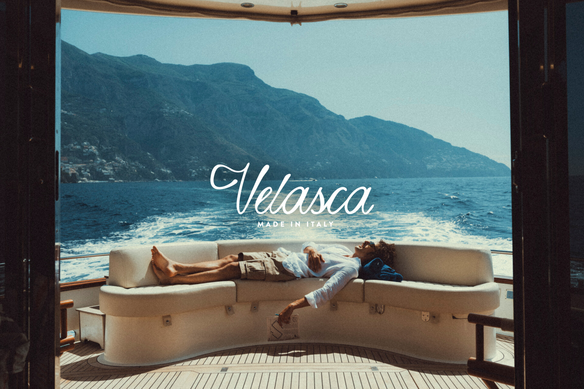 Velasca Made in Italy - Una persona si rilassa su un divano curvo sul retro di uno yacht, con montagne e oceano sullo sfondo. La scritta "Velasca Made in Italy" è sovrapposta al centro dell'immagine.