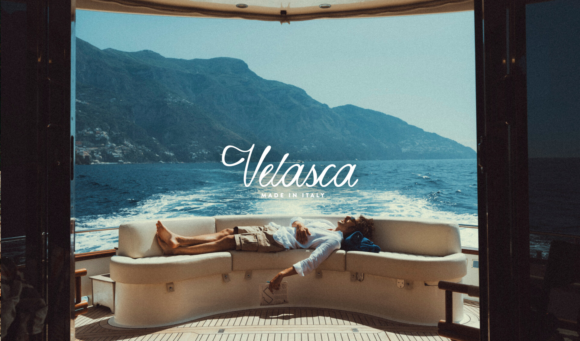 Velasca – Made in Italy - Un uomo è sdraiato su un divano bianco curvo nella parte posteriore di una barca, con lo sguardo rivolto al mare e alle montagne visibili in lontananza; le parole "Velasca" e "Made in Italy" sono sovrapposte in un'elegante calligrafia.