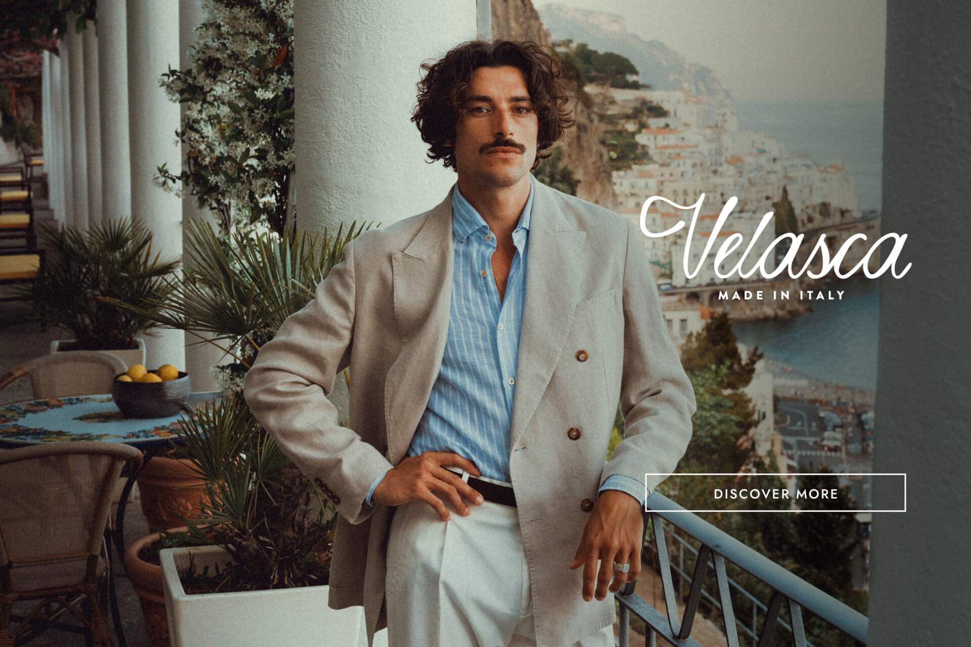 Velasca Made in Italy - Un uomo elegante in abito beige e camicia a righe blu è in piedi su un balcone con vista sulla Costiera Amalfitana. Il testo recita "Velasca Made in Italy" con un pulsante con la scritta "Scopri di più".