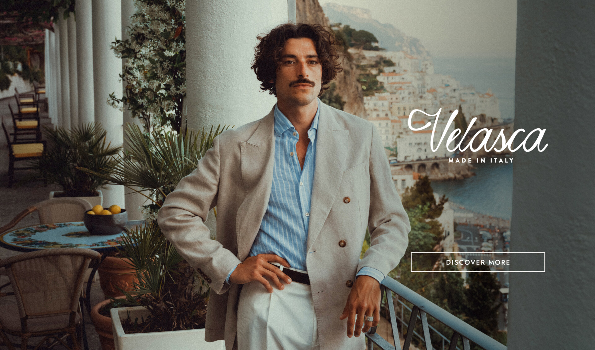 Velasca Made in Italy - Un uomo in abito chiaro e camicia a righe blu è in piedi su un balcone con vista su una città costiera italiana. Nelle vicinanze si trovano tavoli all'aperto e piante rigogliose. Il testo recita "Velasca Made in Italy" con un pulsante "Scopri di più".