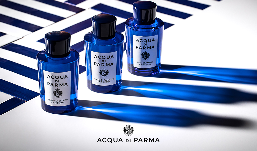 Tre bottiglie blu di Acqua di Parma Cologne - Tre bottiglie blu di Acqua di Parma Cologne sono disposte su una superficie bianca con ombre a strisce blu. Il logo del marchio è visualizzato nella parte inferiore dell'immagine.