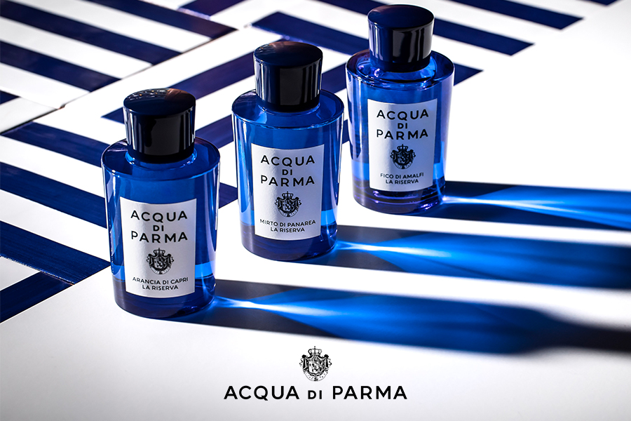 Tre flaconi blu di profumo Acqua di Parma - Tre flaconi blu di profumo Acqua di Parma sono esposti su una superficie bianca con strisce blu diagonali. Le etichette sui flaconi recitano "ARANCIA DI CAPRI", "MIRTO DI PANAREA" e "FICO DI AMALFI". Il logo del marchio Acqua di Parma è posizionato sotto i flaconi.