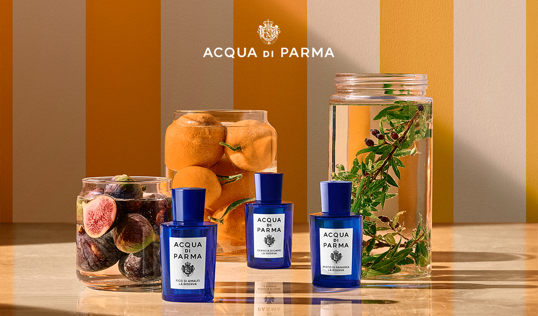 Acqua di Parma - Tre flaconi di profumo blu Acqua di Parma sono esposti su un piano, accanto a barattoli di vetro contenenti arance fresche, fichi e acqua con erbe verdi. Lo sfondo è caratterizzato da strisce verticali gialle e bianche.