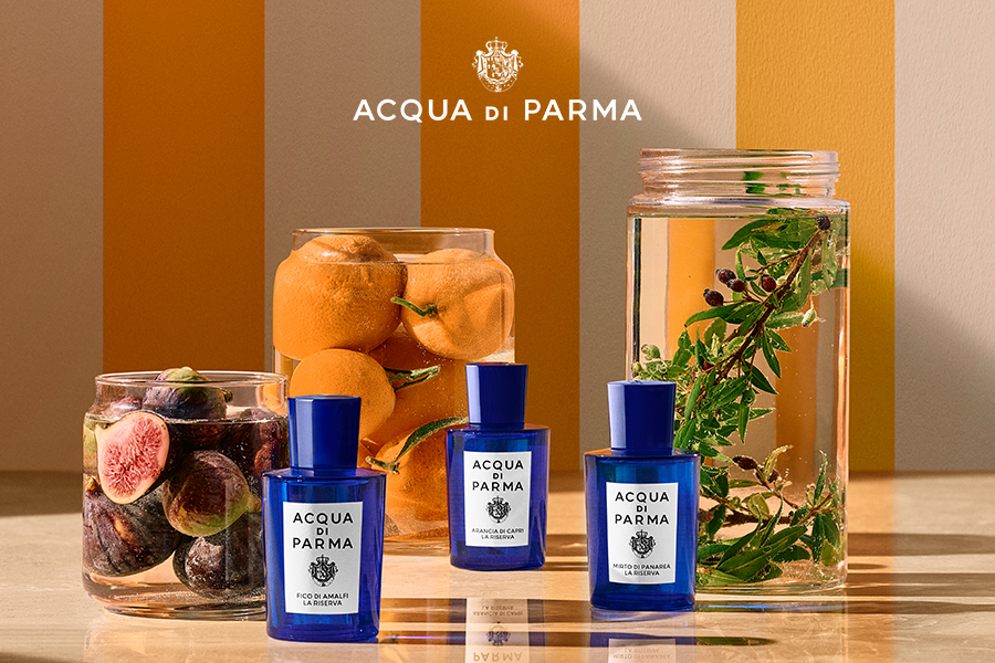 Espositore per profumi Acqua di Parma: tre flaconi di profumo Acqua di Parma blu sono esposti davanti a un barattolo di acqua con erbe aromatiche e un barattolo pieno di fichi e arance, su uno sfondo a righe beige e arancioni. Il logo Acqua di Parma è in alto.