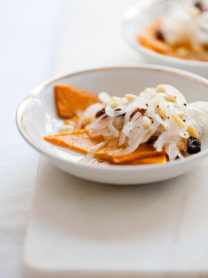 Certainly! Here’s the alt text with the image title at the beginning, following your format: Image Title - A small white plate with thin slices of orange squash topped with pickled onions, raisins, and pine nuts, set on a white surface with another plate blurred in the background.