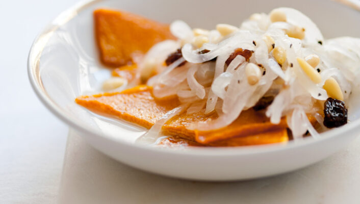 Certo! Ecco il testo alt con il titolo dell'immagine all'inizio: [Titolo immagine] - Un piccolo piatto bianco con sottili fette di zucca arancione arrostita, sormontate da cipolle bianche sottaceto, noci e uvetta, posto su una superficie bianca. Un altro piatto simile appare sfocato sullo sfondo.