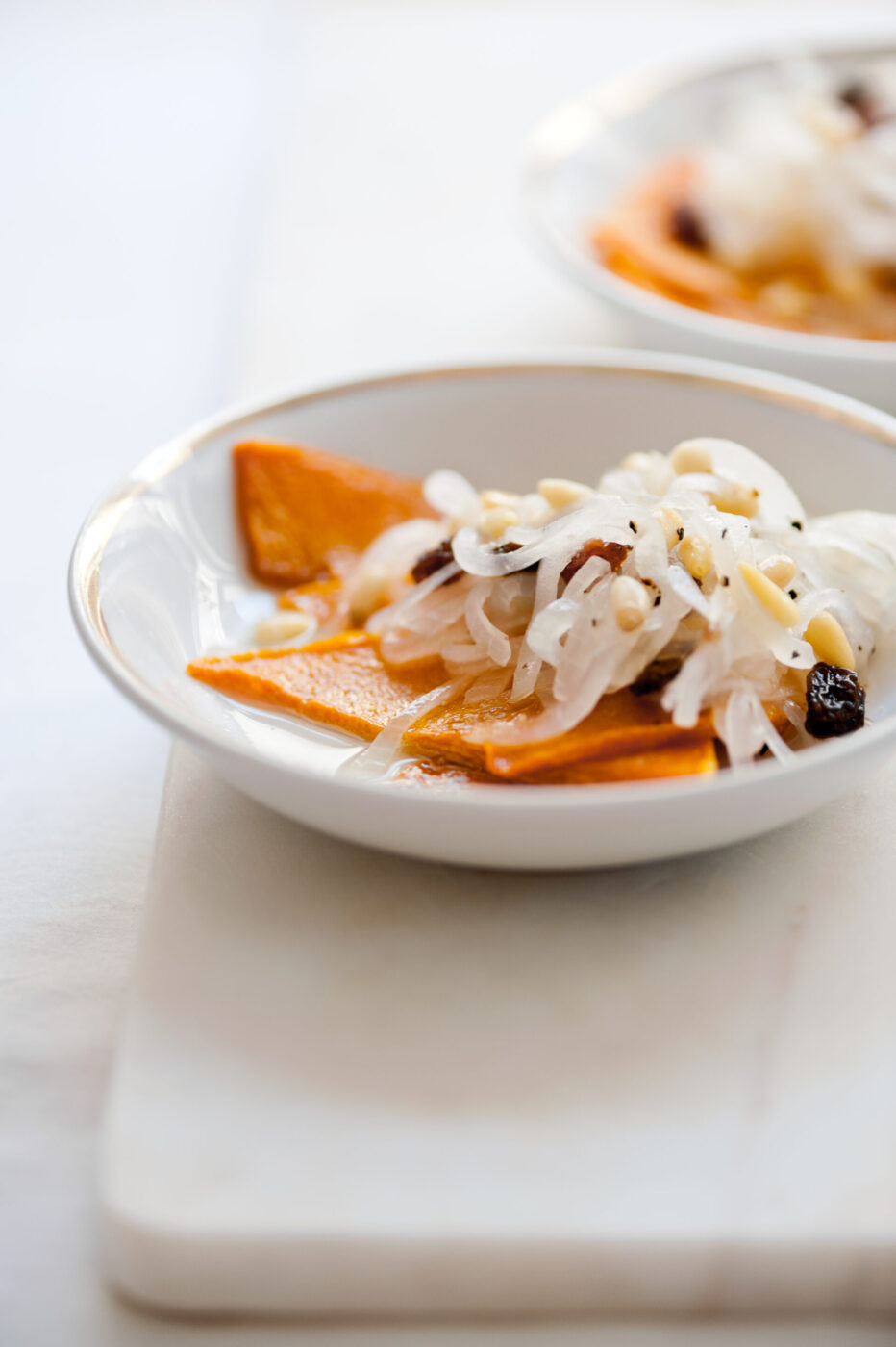Certainly! Here’s the alt text with the image title at the beginning, following your format: Image Title - A small white plate with thin slices of orange squash topped with pickled onions, raisins, and pine nuts, set on a white surface with another plate blurred in the background.