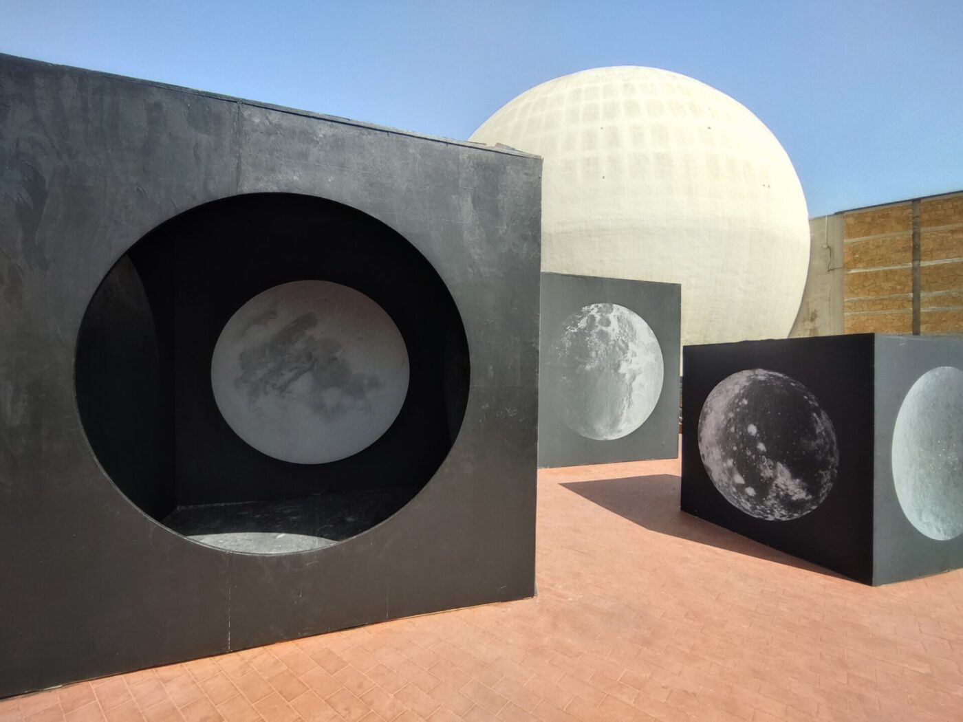 [Grandi cubi neri con ritagli circolari e immagini della luna sono disposti all'aperto su una superficie piastrellata, con una grande struttura a cupola bianca e un cielo azzurro sullo sfondo.