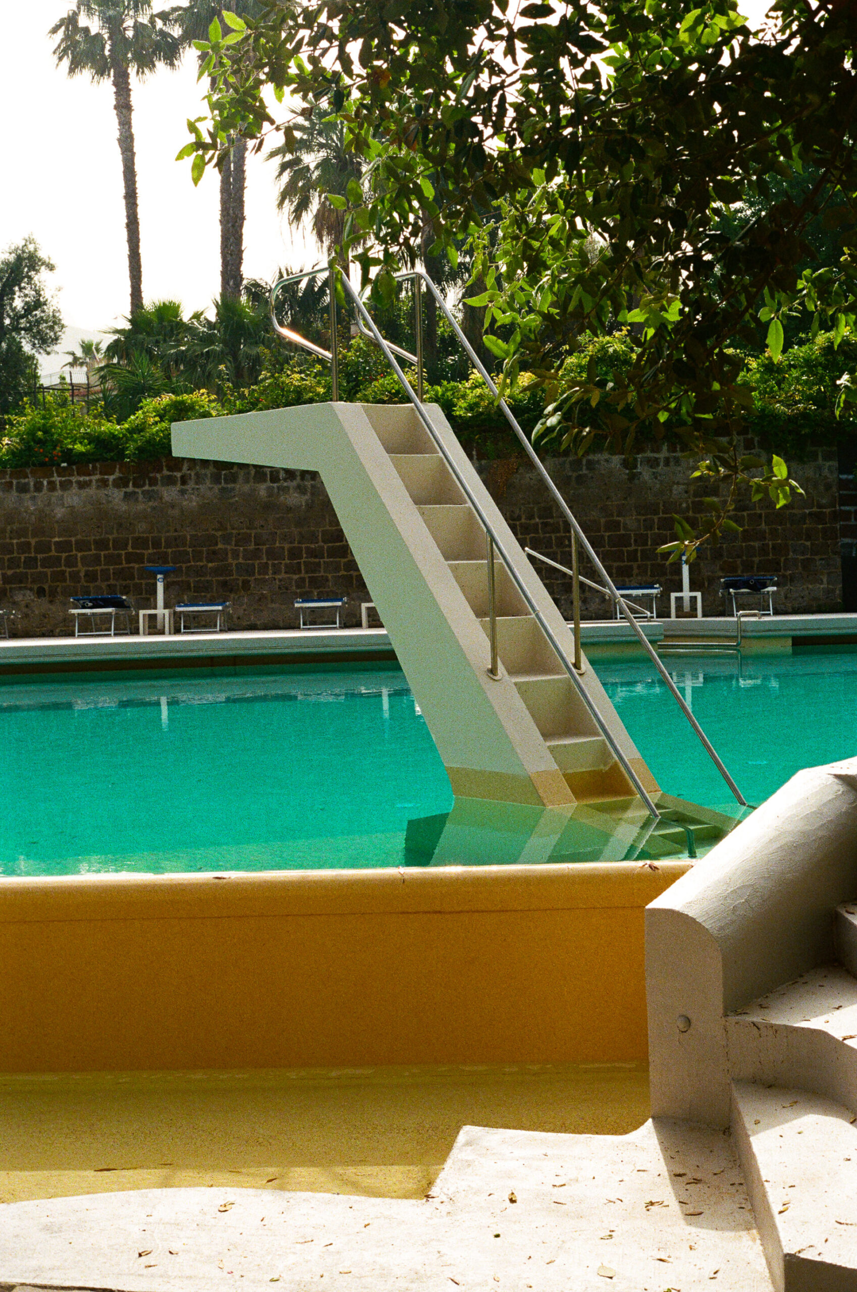A diving board with stairs - A diving board with stairs leads into a clear outdoor swimming pool, surrounded by lounge chairs and palm trees, on a sunny day.