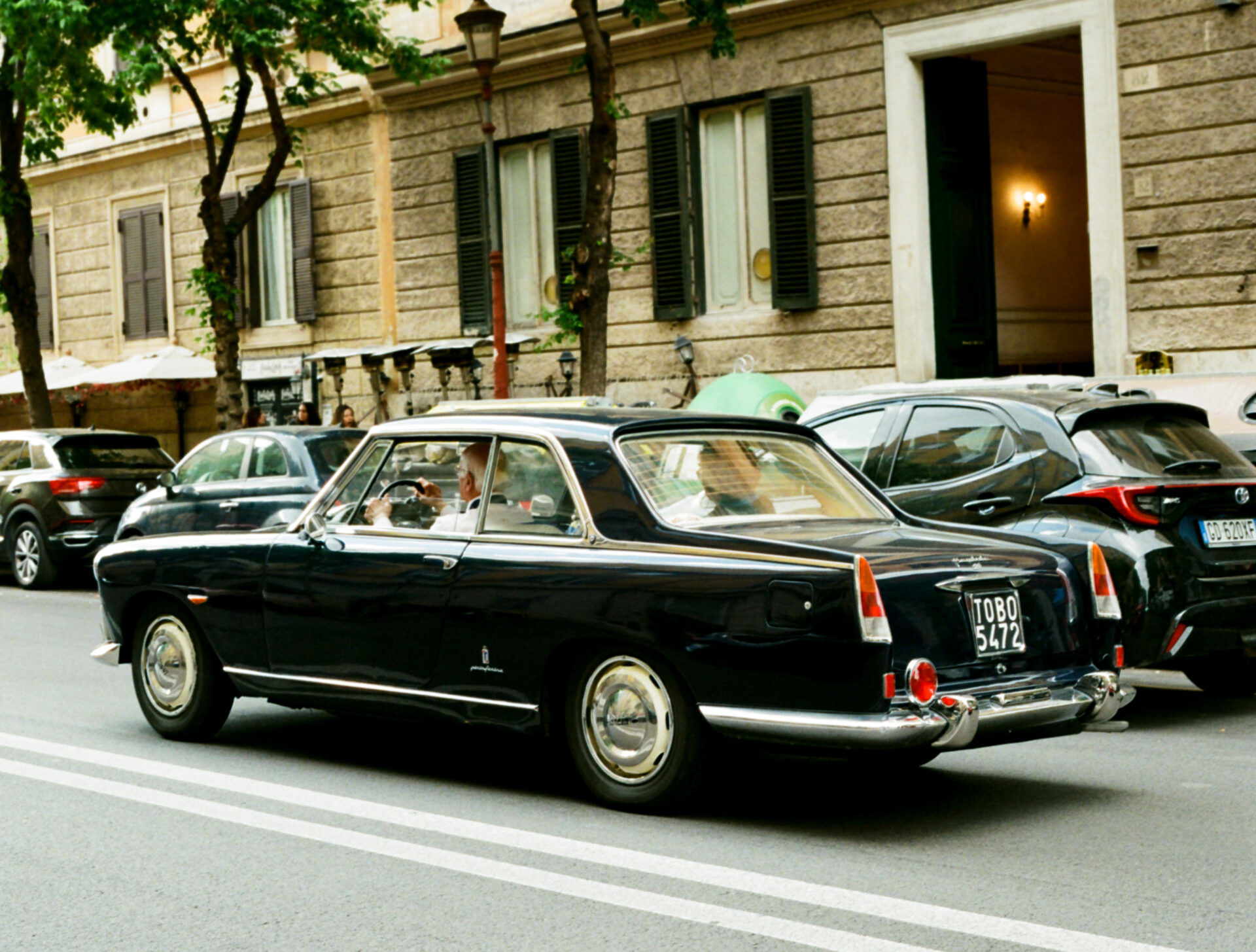 Certamente! Tuttavia, non vedo un titolo esplicito dell'immagine. Se "A classic black car with chrome details drives down a city street" funge da titolo dell'immagine, il testo alt seguendo il vostro formato sarebbe: A classic black car with chrome details drives down a city street - Un'auto nera classica con dettagli cromati percorre una strada cittadina, superando veicoli moderni parcheggiati. La scena è caratterizzata da edifici in pietra con finestre chiuse e un portone ben illuminato sullo sfondo. Se si desidera un'altra frase come titolo, si prega di specificarlo!.
