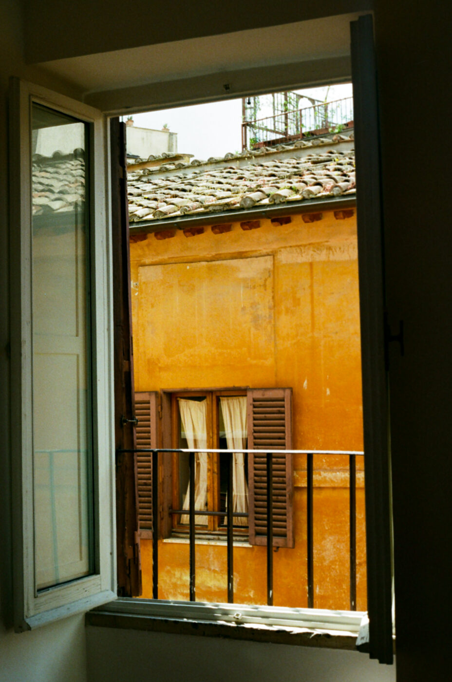 Una finestra aperta con persiane bianche e azzurre rivela la vista di un edificio giallo-arancio con tetto di tegole, persiane marroni e tende bianche, visto attraverso una stretta ringhiera del balcone. (Nota: non è stato fornito il titolo dell'immagine. Se si fornisce il titolo di un'immagine, lo inserirò all'inizio come indicato).