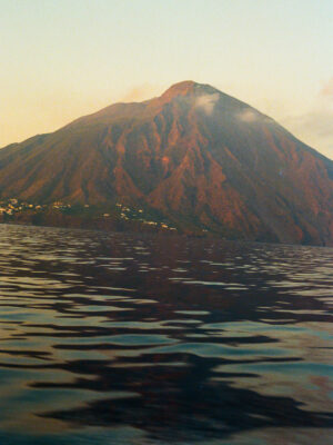 Sunset Volcano Reflection - A large volcanic mountain rises above calm, reflective water at sunset, with warm light highlighting its slopes and a few small clouds near the peak.