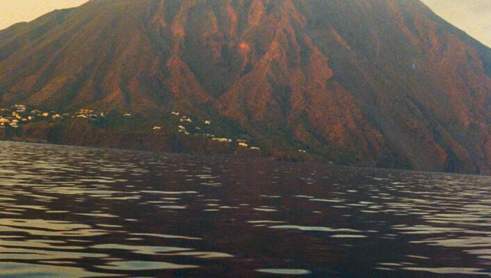 [Una grande montagna si erge su acque calme e increspate al tramonto. La montagna ha pendici bruno-rossastre con deboli tracce di vegetazione e piccoli edifici bianchi sono sparsi lungo la sua base.