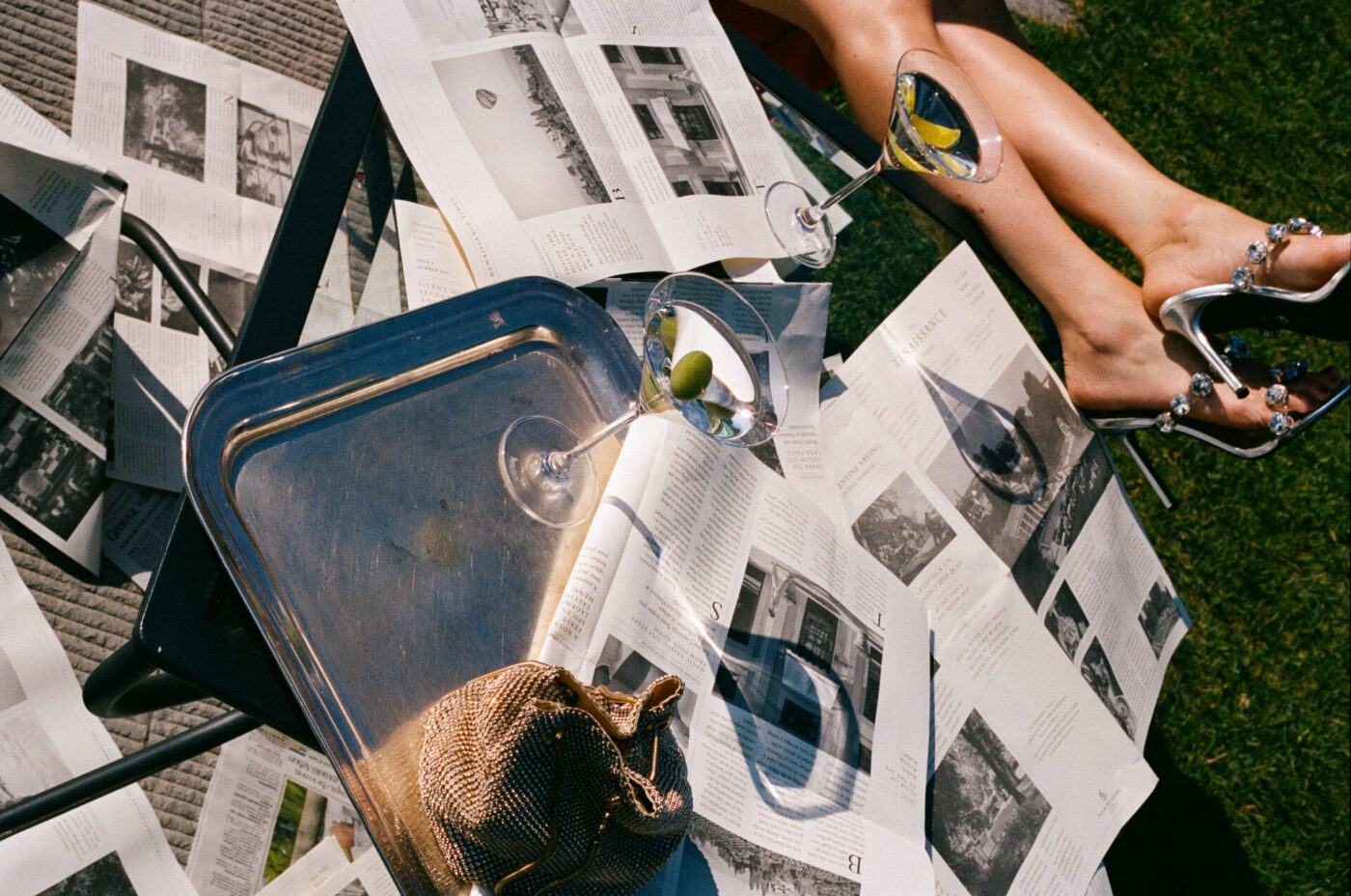 A person’s legs with jeweled sandals rest on a chair surrounded by scattered open magazines, a metallic purse, and two martini glasses, one on a silver tray, all arranged on grass in bright sunlight.