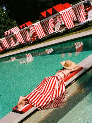 Summer Poolside Relaxation - A person wearing a red and white striped dress and a straw hat lounges on a red pool float, with matching red and white striped cabanas and sun loungers in the background by a clear swimming pool. Let me know if you’d like to use a different title!.