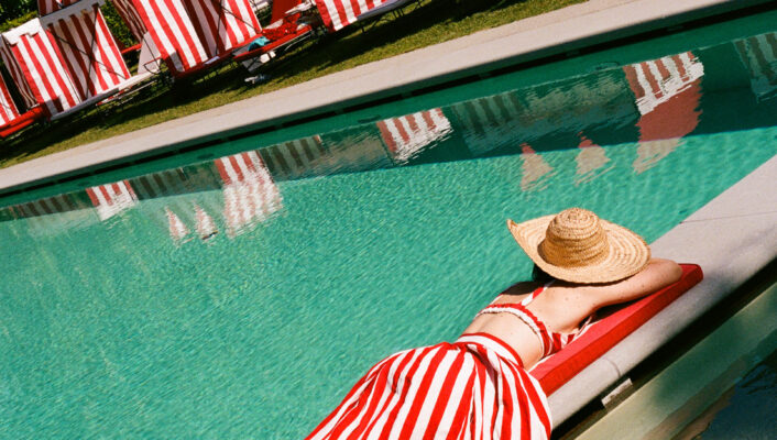 Ecco il testo alt seguendo le tue istruzioni: Una persona che indossa un vestito a righe bianche e rosse e un cappello di paglia si sdraia a bordo piscina, circondata da cabanas a righe bianche e rosse in una giornata di sole. Se è presente un titolo dell'immagine, vi prego di fornirlo, in modo che possa aggiungerlo al testo alt come richiesto. Se non c'è un titolo dell'immagine, il testo alt di cui sopra è corretto.