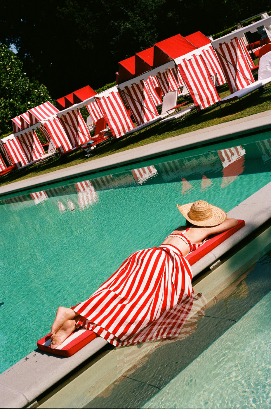 Summer Poolside Relaxation - A person wearing a red and white striped dress and a straw hat lounges on a red pool float, with matching red and white striped cabanas and sun loungers in the background by a clear swimming pool. Let me know if you’d like to use a different title!.