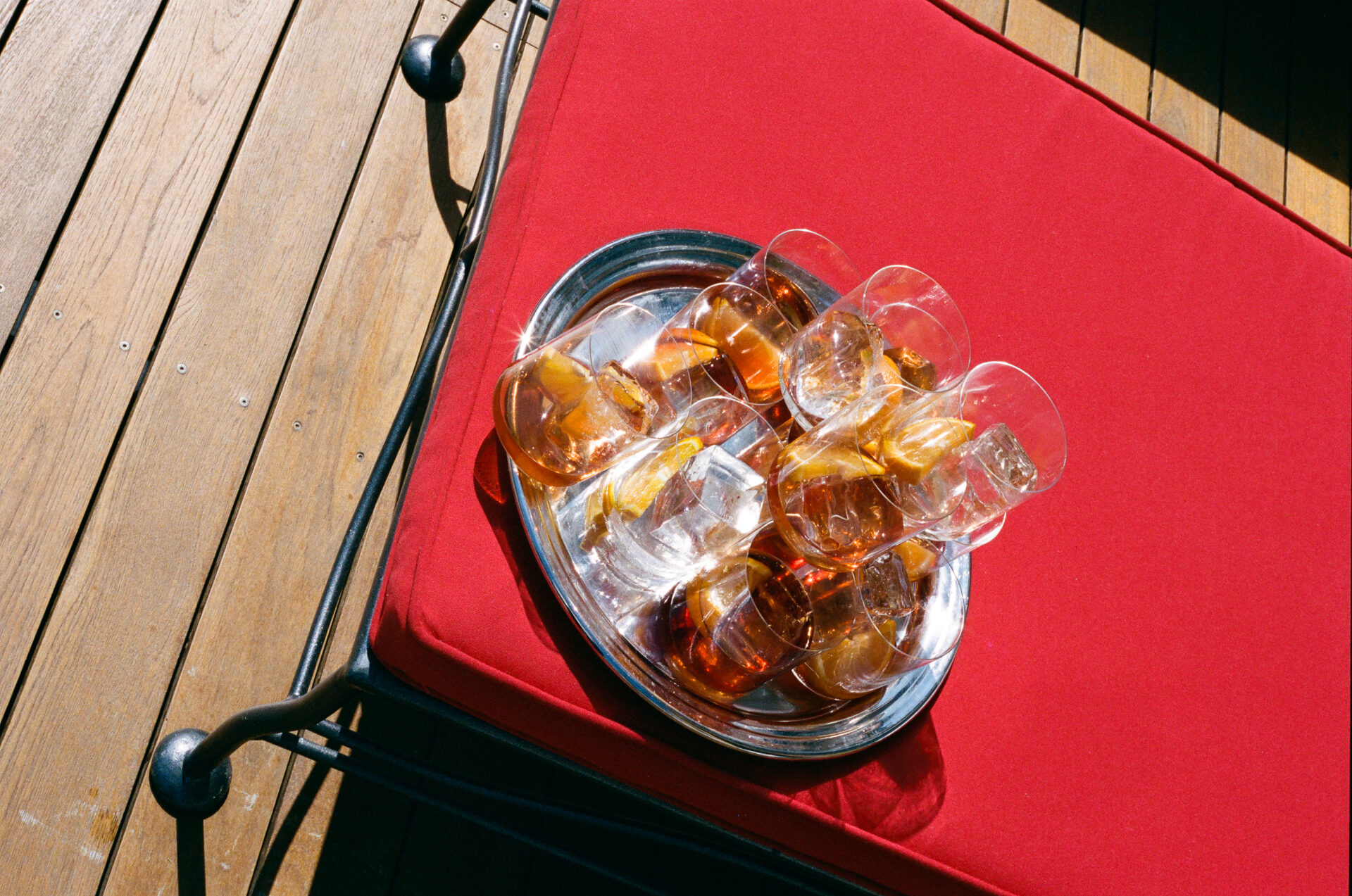 A metal tray with drinks on chair - A metal tray with several glasses of drinks, some clear and some amber-colored with ice and lemon slices, sits on a red cushioned chair on a wooden deck in sunlight.