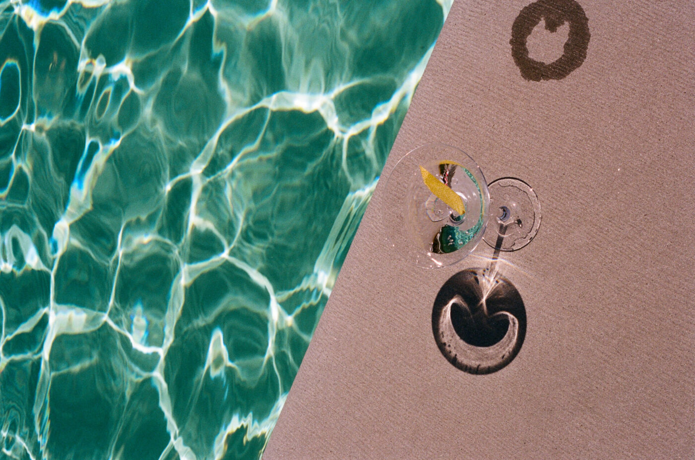 A glass of water with a lemon slice sits on the edge of a swimming pool, casting shadows and a ring mark on the poolside surface next to shimmering turquoise water.