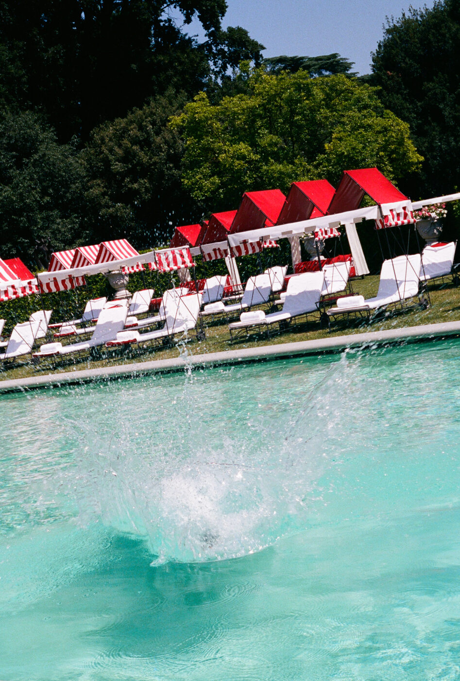 A large splash is seen in a swimming pool, suggesting someone just jumped in. In the background, there are red and white striped cabanas and lounge chairs on a sunny day.