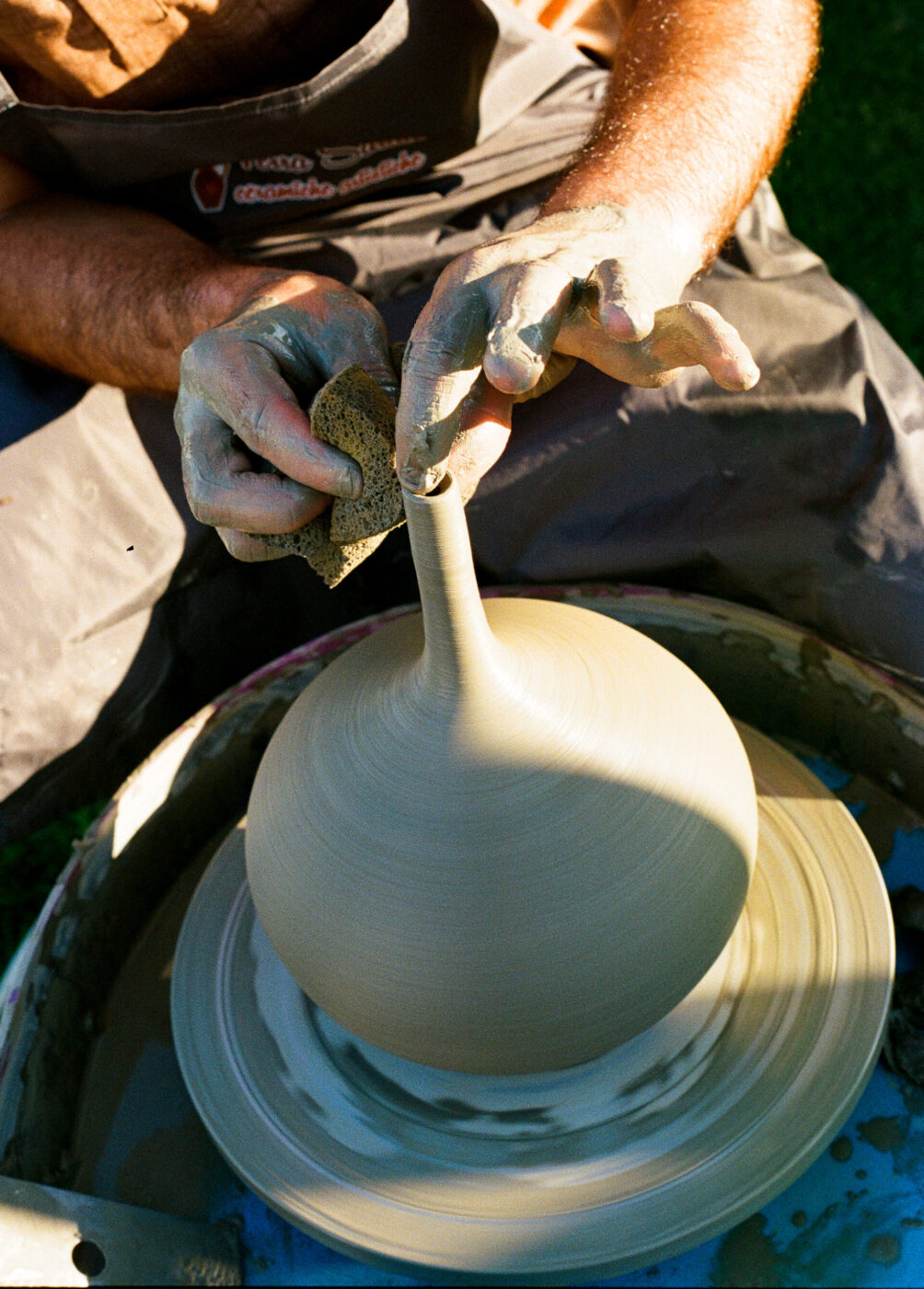 Mani che modellano un vaso di argilla - Mani che modellano un vaso di argilla alto e dal collo stretto su un tornio, usando una spugna per lisciare; il vasaio indossa un grembiule grigio e ha l'argilla sulle dita.